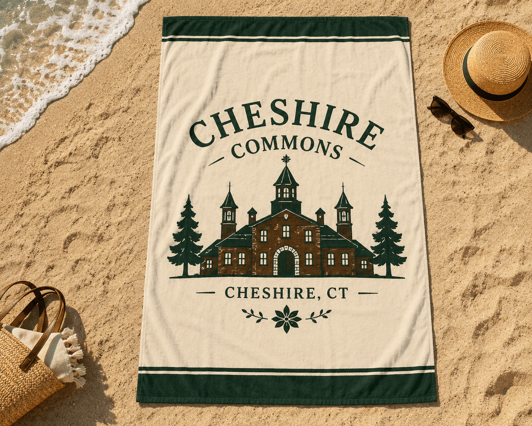 Cheshire Commons beach towel laid out on sand, crest and arched wordmark centered